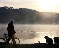 SILHUETA DE UM CICLISTA PASSANDO AO LADO DE CAPIVARAS NO PARQUE BARIGUI COM O SOL NASCENDO AO FUNDO